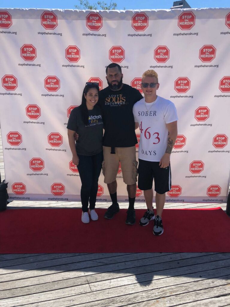 Three people stand on a red carpet in front of a Stop Heroin step-and-repeat banner. One person wears a Sober 463 Days shirt, another wears a dark Souls shirt, and the third wears a dark Alcohol shirt.