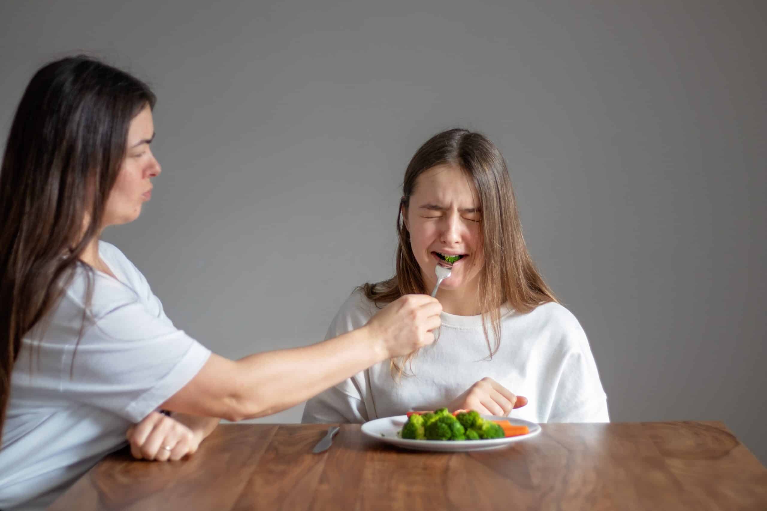 Mother forces her crying teen daughter to eat healthy food feeding her broccoli with fork. Food waste. No vegan diet concept. No choice