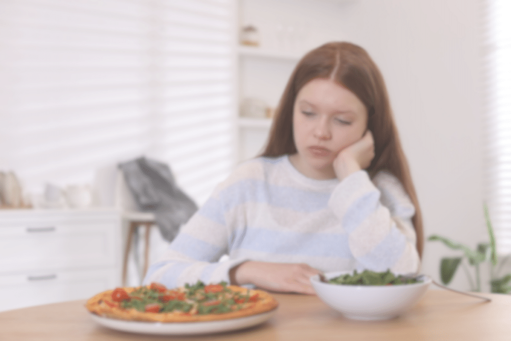 A girl with long brown hair sits at a table, looking bored or uninterested, with a bowl of salad and a vegetable pizza in front of her in a bright, modern kitchen.