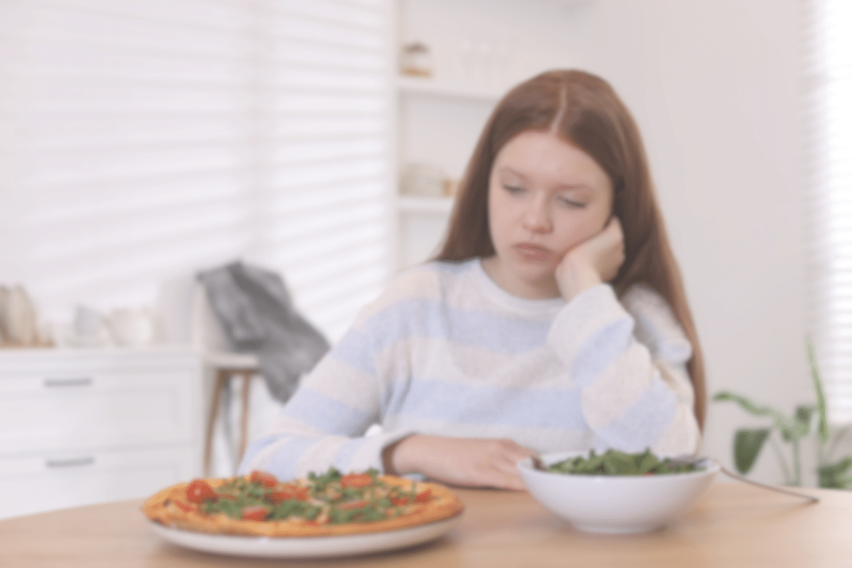 A girl with long brown hair sits at a table, looking bored or uninterested, with a bowl of salad and a vegetable pizza in front of her in a bright, modern kitchen.