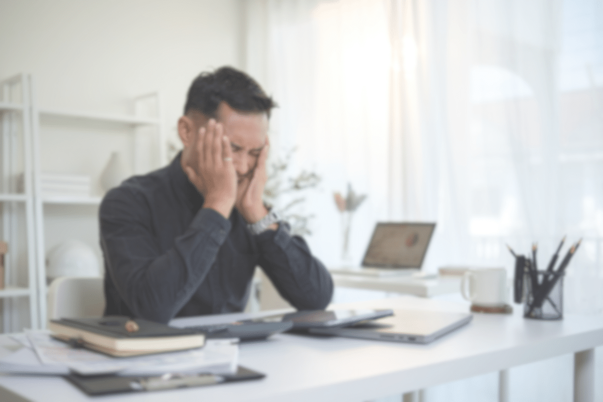 A man sits at a desk with his head in his hands, appearing stressed or overwhelmed. The desk is cluttered with notebooks, papers, a laptop, and a coffee mug in a bright, modern office. The image is blurred.