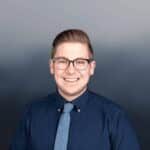 A young man with short, light brown hair and glasses, wearing a navy blue button-up shirt and a light blue tie, smiles at the camera against a gradient gray background.