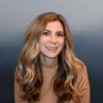 Smiling woman with long, wavy blonde hair wearing a tan sweater, posing in front of a gradient blue background.