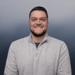 A man with short brown hair and a trimmed beard smiles at the camera. He is wearing a light gray quarter-zip sweater and stands in front of a gradient blue-gray background.