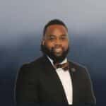 A man in a black suit with a bow tie and a pin on his lapel smiles at the camera against a gradient blue-gray background.