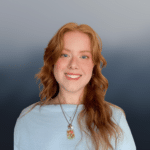 A young woman with long, wavy red hair smiles at the camera. She wears a light blue top and two necklaces, one with a colorful pendant. The background is a smooth blue-gray gradient.