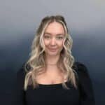 A woman with long, wavy blonde hair and blue eyes is wearing a black top and a gold necklace, standing against a blurred gradient blue and gray background. She is smiling softly at the camera.