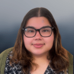 A woman with glasses and shoulder-length brown hair smiles softly, wearing a patterned top and green sweater, against a blurred blue-gray background.
