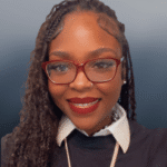 Smiling woman with long braided hair, wearing red glasses, red lipstick, and a black shirt over a white collared top, against a plain blurred background.