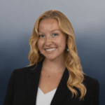 A woman with long wavy blonde hair, wearing a black blazer over a white top, smiles at the camera against a gradient blue background.