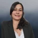 A woman with long dark hair, wearing a black blazer and white lace-trim top, smiles at the camera against a soft gradient blue-gray background.