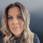 A person with long wavy light brown hair and light makeup smiles slightly while wearing a dark top, set against a soft blue gradient background.