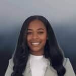 A woman with long, wavy dark hair smiles at the camera. She wears a light-colored blazer over a white top. The background is a smooth, gradient blend of blue and gray tones.