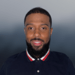 A man with short hair and a beard, smiling, wears a dark polo shirt with red, white, and blue striped collar, standing against a blurred gray background.