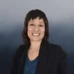 A woman with short brown hair and bangs smiles at the camera. She is wearing a dark blazer over a blue top, with a blurred gray and blue background.