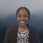 A woman with braided hair smiles at the camera, wearing a black blazer and a patterned top with a pendant necklace, against a blue gradient background.