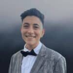 A person with short dark hair, wearing a gray plaid suit jacket, white shirt, black bow tie, and hoop earring, smiles at the camera in front of a blurred, gradient gray background.