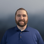 A man with a beard and long hair pulled back, wearing a blue polo shirt, stands smiling in front of a blurred gray-blue background.