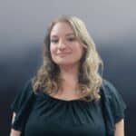 A woman with wavy, shoulder-length blonde hair is standing against a gradient gray background. She is wearing a black top and a necklace, and she has a slight smile.