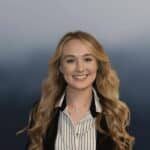 A young woman with long, wavy blonde hair smiles at the camera. She is wearing a black blazer over a white shirt with black stripes, and the background is a smooth gradient of blue tones.