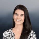 A woman with long dark hair, wearing a black and white patterned top, smiles at the camera against a smooth, dark gradient background.