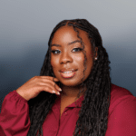 A woman with long braided hair and a burgundy blouse smiles gently, resting her hand near her chin, posed against a soft, gradient gray background.