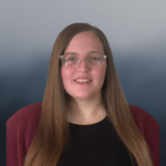 Woman with long straight brown hair, wearing clear glasses, a black top, and a maroon cardigan, smiling at the camera. The background is a smooth gradient of blue and gray tones.