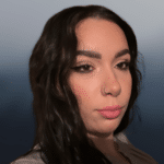 A person with long wavy dark hair, thick eyebrows, and winged eyeliner looks at the camera against a blurred blue and gray background. They have a nose ring and neutral makeup.