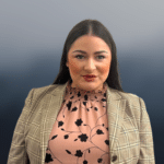 Woman with long dark hair wearing a plaid blazer over a pink floral blouse, standing in front of a gradient gray background.