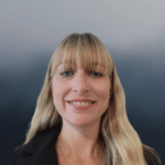 A woman with long blonde hair and bangs, smiling at the camera. She is wearing a black blazer and is set against a gradient blue-grey background.
