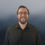 A man with glasses, a beard, and short dark hair smiles while wearing a black button-up shirt. The background is blurred with blue and gray tones.