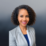 A smiling woman with curly hair wearing a gray blazer, white blouse, and blue top, with gold hoop earrings, poses in front of a neutral gray background.
