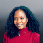 A woman with curly black hair smiles gently, wearing a red turtleneck sweater and a necklace, against a soft gray background.