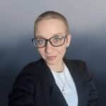 A person with short hair, glasses, and light skin is wearing a black blazer over a white top and a necklace, smiling at the camera against a gradient grey background.