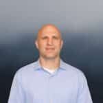 A bald man wearing a light blue button-up shirt stands in front of a blurred, dark blue and gray background, facing the camera with a neutral expression.