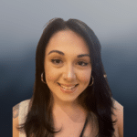 A woman with long dark hair, hoop earrings, and a nose ring smiles at the camera against a blurred grey-blue background.