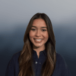 A young woman with long brown hair and a dark shirt smiles in front of a blurred blue and gray gradient background.