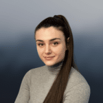 A young woman with long brown hair in a ponytail, wearing a gray turtleneck, smiles softly at the camera against a smooth, blurred blue background.