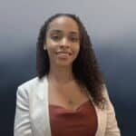 A woman with curly hair wearing a white blazer over a red top smiles at the camera, standing in front of a blurred blue and gray background.