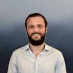 A man with a beard and mustache, wearing a light blue checked shirt, stands and smiles at the camera. The background is a gradient of dark blue and gray tones.