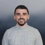 A man with short dark hair and a beard is smiling at the camera, wearing a light gray knit sweater. The background is a gradient of dark and light blue tones.