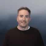 A man with short brown hair and light stubble, wearing a black sweater over a white shirt, stands against a blue gradient background, smiling slightly at the camera.