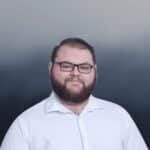 A bearded man with short brown hair wears black glasses and a white collared shirt, smiling slightly. The background is a blurred gradient of dark blue and gray tones.